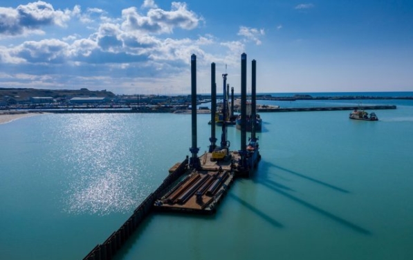 A barge with construction equipment sits on tranquil water under a blue sky. The barge is close to shore.