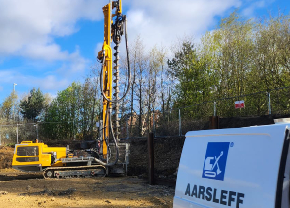 A yellow Aarsleff piling rig stands tall on a construction site. A white van with the Aarsleff company logo is seen on the right.