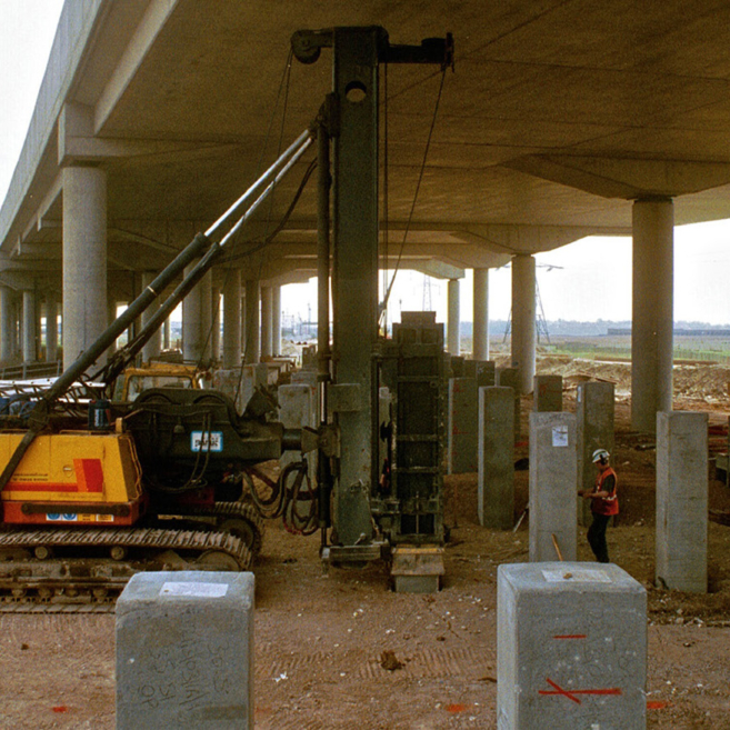 Piling machinery operates under a highway overpass. Many precast concrete piles are present with a worker in safety PPE standing nearby.