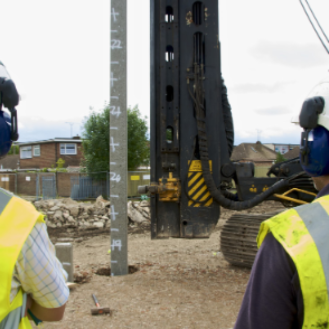 Two workers wearing Aarsleff vests and white hard hats look at a pile driver in use.