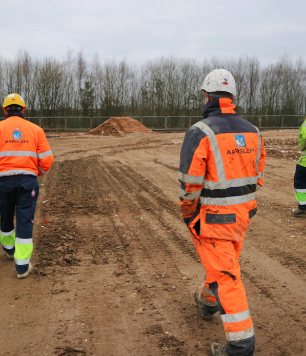 Workers In PPE Walking Towards A Site. Two Workers Are Wearing A Orange Aarsleff Jacket.