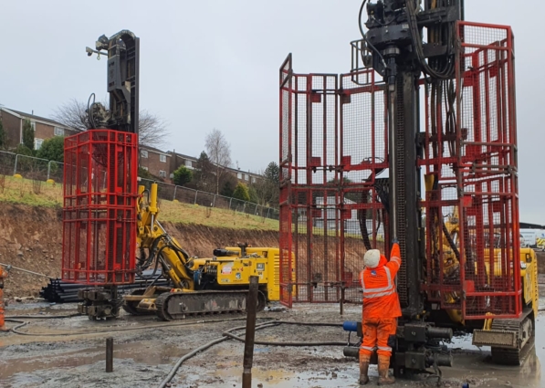 Two yellow drilling rigs sit a muddy construction site. A worker in an orange safety gear works near one of the rigs.