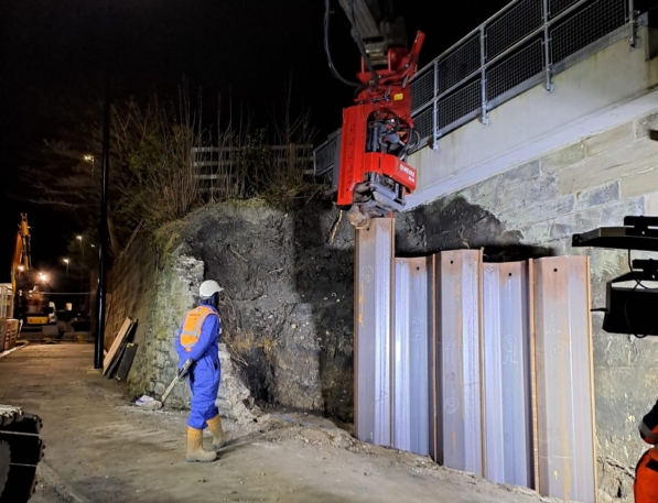 Steel sheet piles being installed by a rig at night. An Aarsleff worker in safety gear observes nearby.