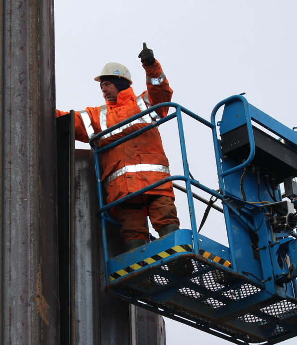A worker in PPE stands in a elevated lift, checks the installation of a sheet pile, gesturing with one hand.