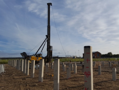 A wide shot of a construction site with square precast concrete piles placed into ground in a grid pattern. A pile driving rig is visible in the background.