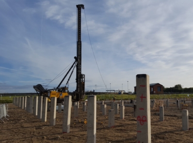 A wide shot of a construction site with square precast concrete piles placed into ground in a grid pattern. A pile driving rig is visible in the background.