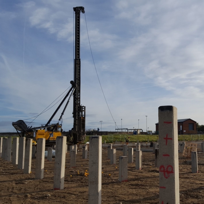A wide shot of a construction site with square precast concrete piles placed into ground in a grid pattern. A pile driving rig is visible in the background.