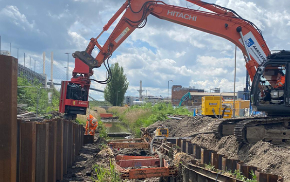 An Aarsleff plant operator installed sheet piles with a large Hitachi excavator. Another construction worker is visible near the sheet piled wall.