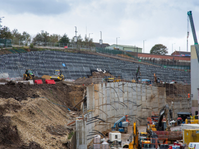 A soil wall being constructed under a cloudy sky. Construction workers and heavy machinery is visible.