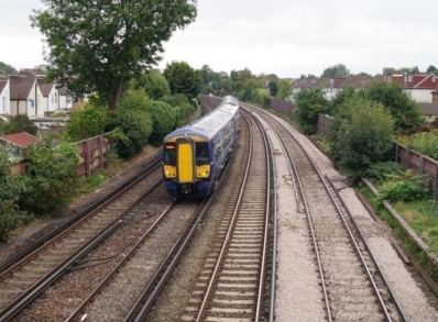 A blue and yellow train moves along railway tracks. Houses and trees is visible on both side of the railway.