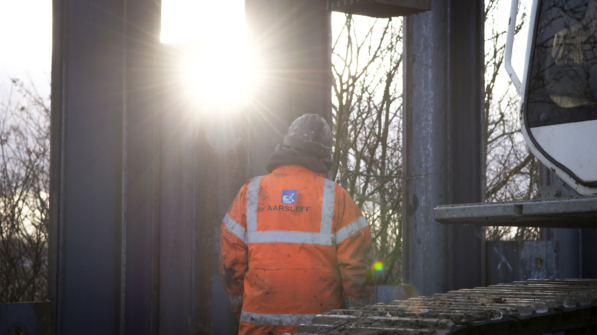 A worker wearing a orange Aarsleff hi-vis jacket and PPE stands in front of sheet piles being installed. The sun shines brightly between the sheet piles.