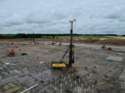 A piling rig positioned in the centre on a piling mat and surrounded by concrete piles. Workers and other heavy machinery are visible.