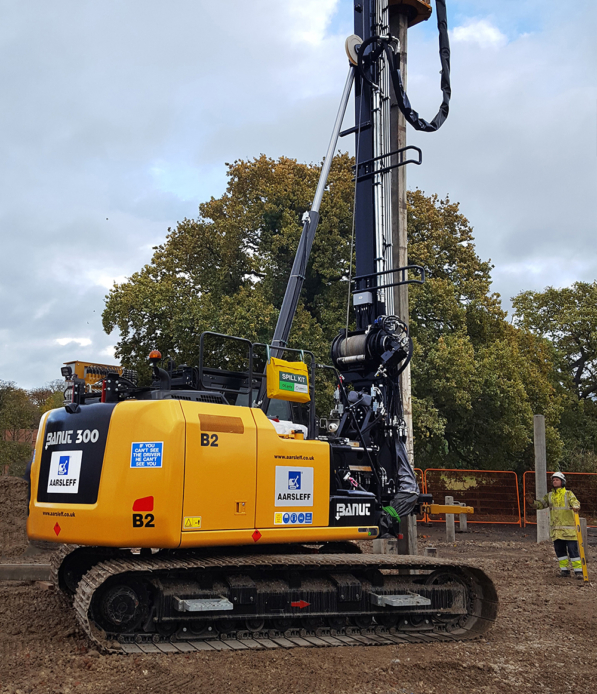 Aarsleff's yellow Banut 300 piling rig in operation. A worker stands nearby to guide the rig operator.