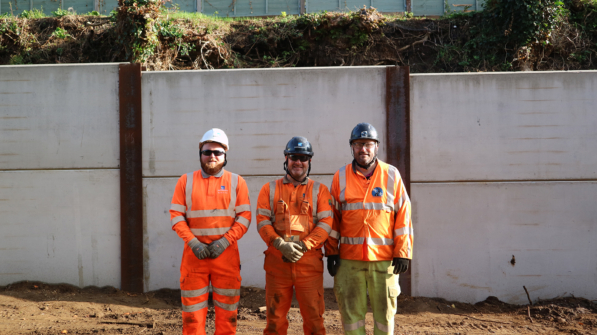 Three Aarsleff Workers Standing In Front Of A King Post Wall. The Workers Are Wearing Orange Hi-Vis Jackets And Hard Hats.