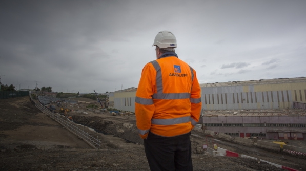 An employee wearing an orange Aarsleff hi-vis jacket and white hard hat stands on a construction site.
