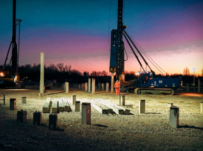 A groundwork project being performed at sunset, illuminated by floodlights. A construction worker and a blue piling rig in operation is visible, surrounded by concrete piles.