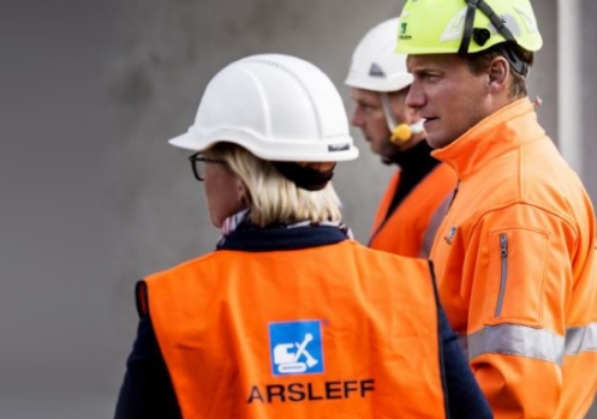 Three construction workers wearing hard hats are looking forward. The female worker in front is wearing an orange vest with the Aarsleff logo on the back of it.