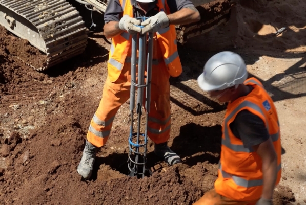 Two workers in bright orange vests and PPE install a steel pile.