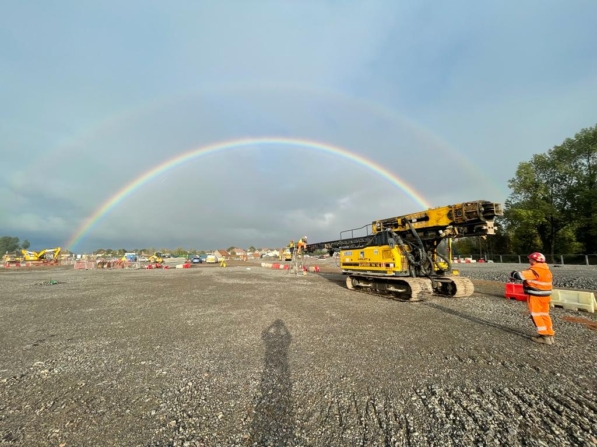 Double rainbow arching over a construction site with Aarsleff's piling rig and a worker in the foreground.