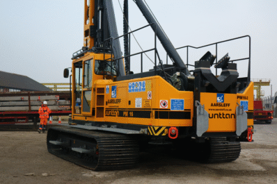 Close-up of Aarsleff's yellow Junttan PM 16 pile driving rig. A worker in safety gear guides the rig operator.