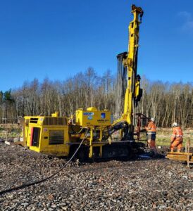 Two Aarsleff workers providing drilling and grouting services with a yellow drilling rig under a clear, blue sky.