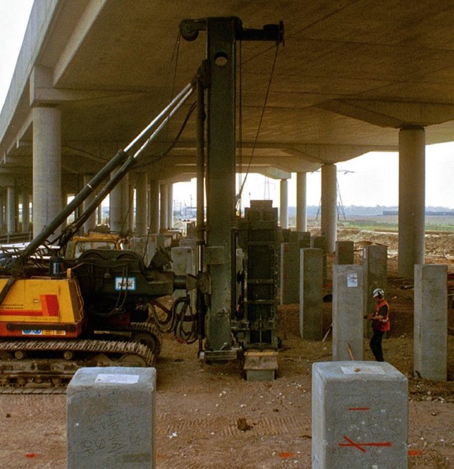 Piling machinery operates under a highway overpass. Many precast concrete piles are present with a worker in safety PPE standing nearby.
