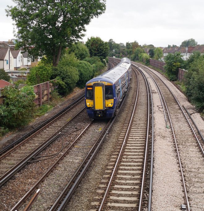 A blue and yellow train moves along railway tracks. Houses and trees is visible on both side of the railway.