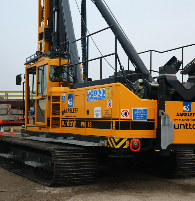 Close-up of Aarsleff's yellow Junttan PM 16 pile driving rig. A worker in safety gear guides the rig operator.