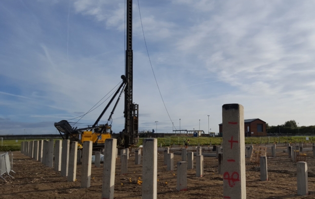 A wide shot of a construction site with square precast concrete piles placed into ground in a grid pattern. A pile driving rig is visible in the background.