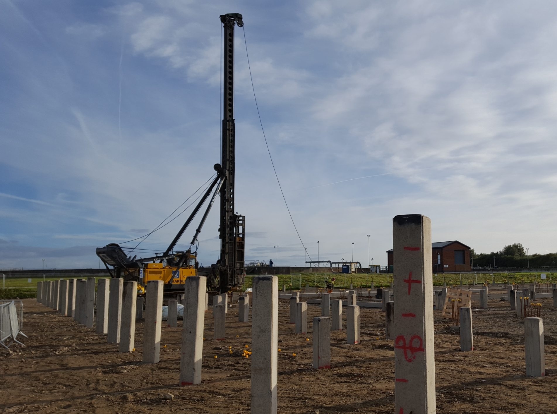 A wide shot of a construction site with square precast concrete piles placed into ground in a grid pattern. A pile driving rig is visible in the background.