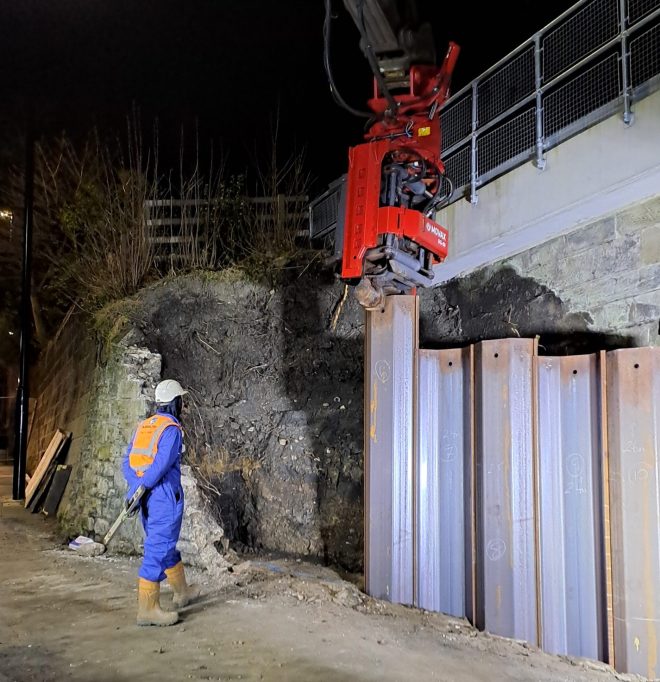 Steel sheet piles being installed by a rig at night. An Aarsleff worker in safety gear observes nearby.