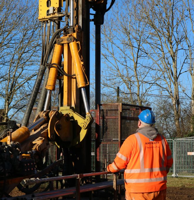 A worker in a orange Aarsleff jacket and blue hard hat stands near a yellow piling machine under a blue sky.