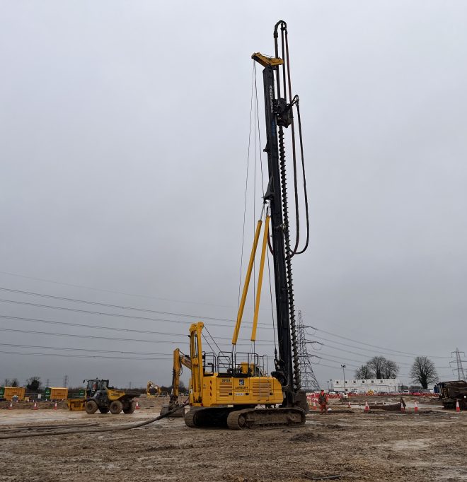 Yellow piling rig sits on a muddy site under a cloudy sky. Other heavy machinery is visible in the background.