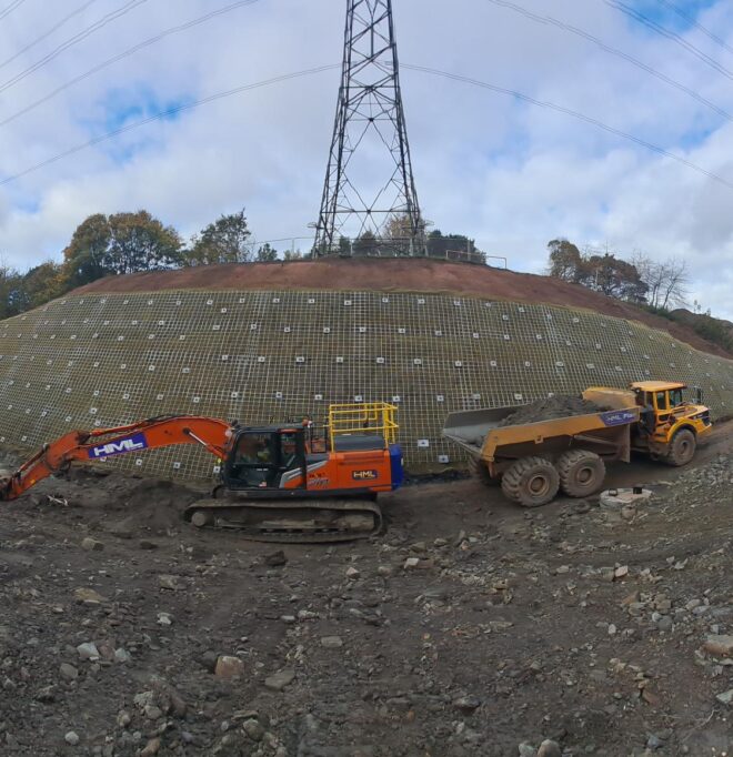 Wide-angle shot of soil nails installed on a slope. Heavy machinery is parked in front and a transmission tower is visible on top of the slope.